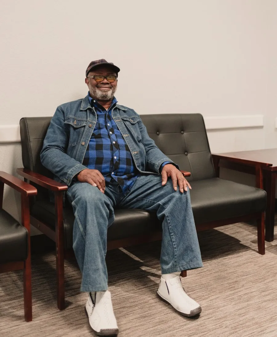 Smiling older man wearing a denim jacket, blue plaid shirt, jeans, and white shoes sitting on a black leather bench in a waiting room.