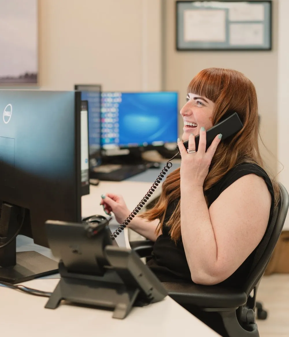Smiling woman talking on a desk phone while seated at a computer workstation in an office.