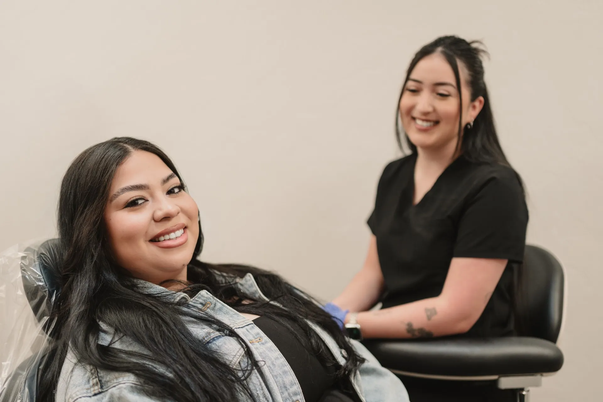 Smiling woman in a denim jacket reclined in a dental chair with a dental hygienist in black scrubs smiling in the background.