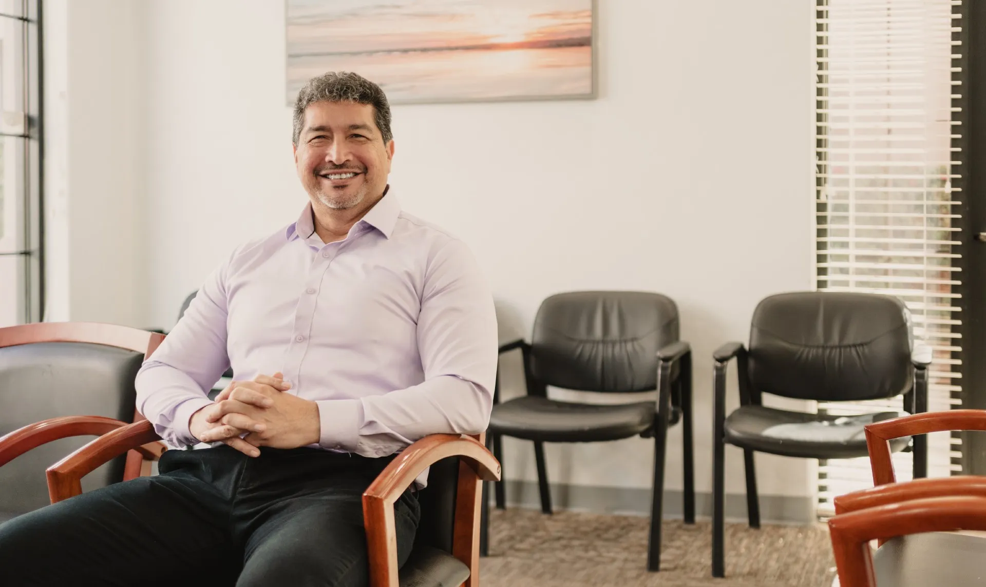 Smiling man in a light purple shirt sitting on a wooden armchair in a waiting room with black chairs and a sunset painting.