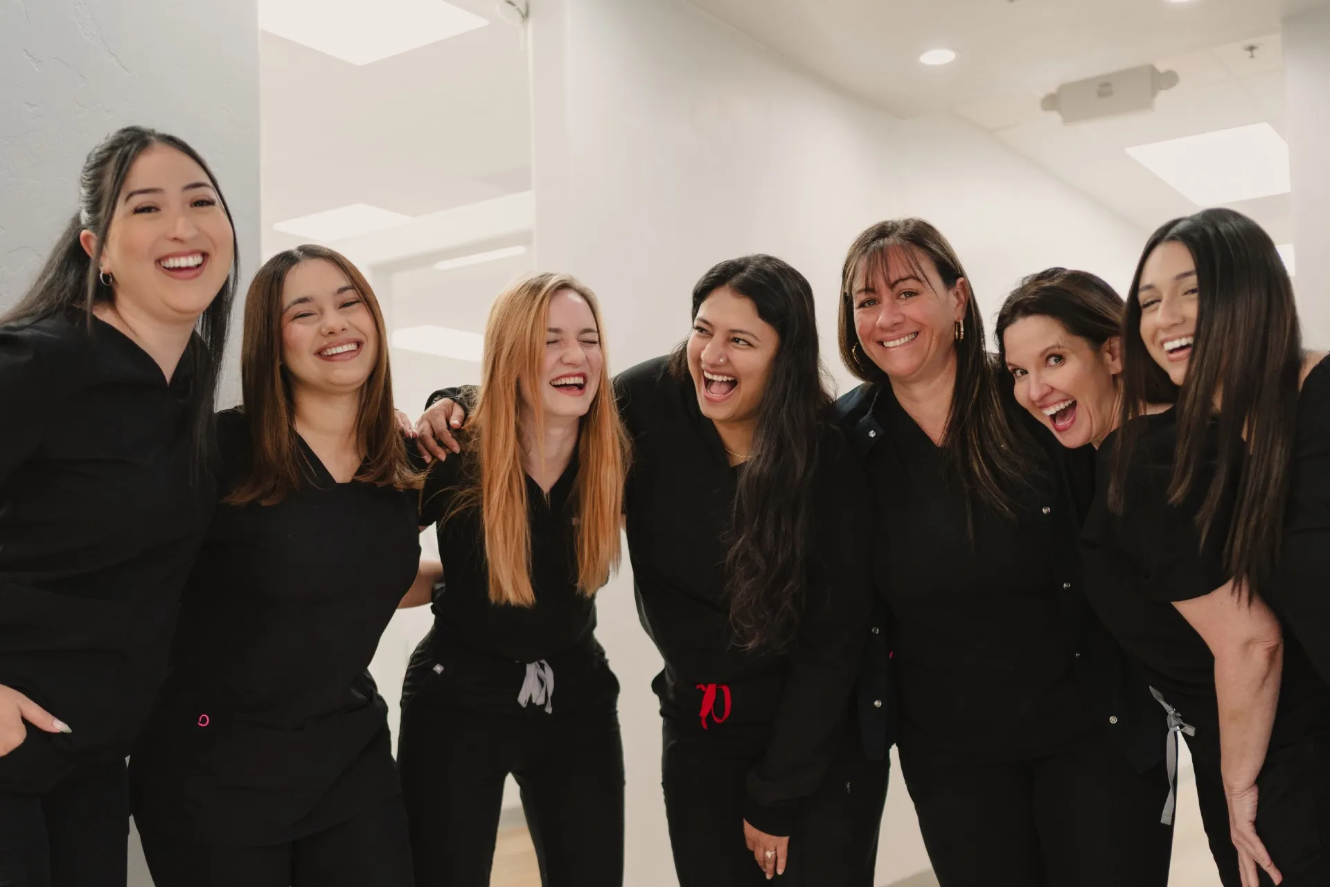 Group of seven women in black scrubs smiling and laughing together indoors.