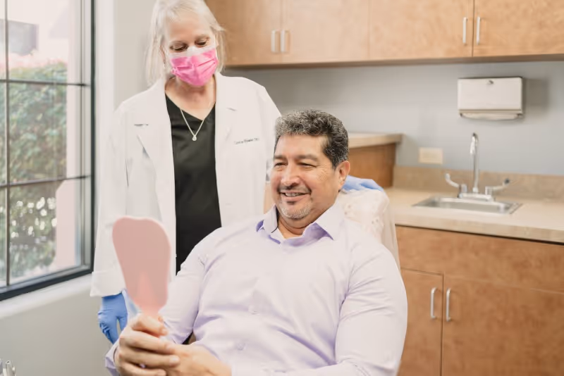 Smiling man in dental chair holding mirror, with masked female dentist standing behind him.