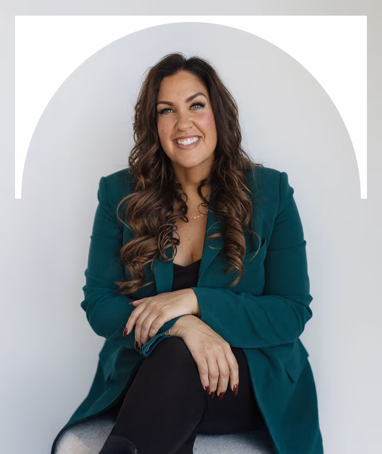 Woman with long curly hair wearing a green blazer sitting with her hands folded and smiling.