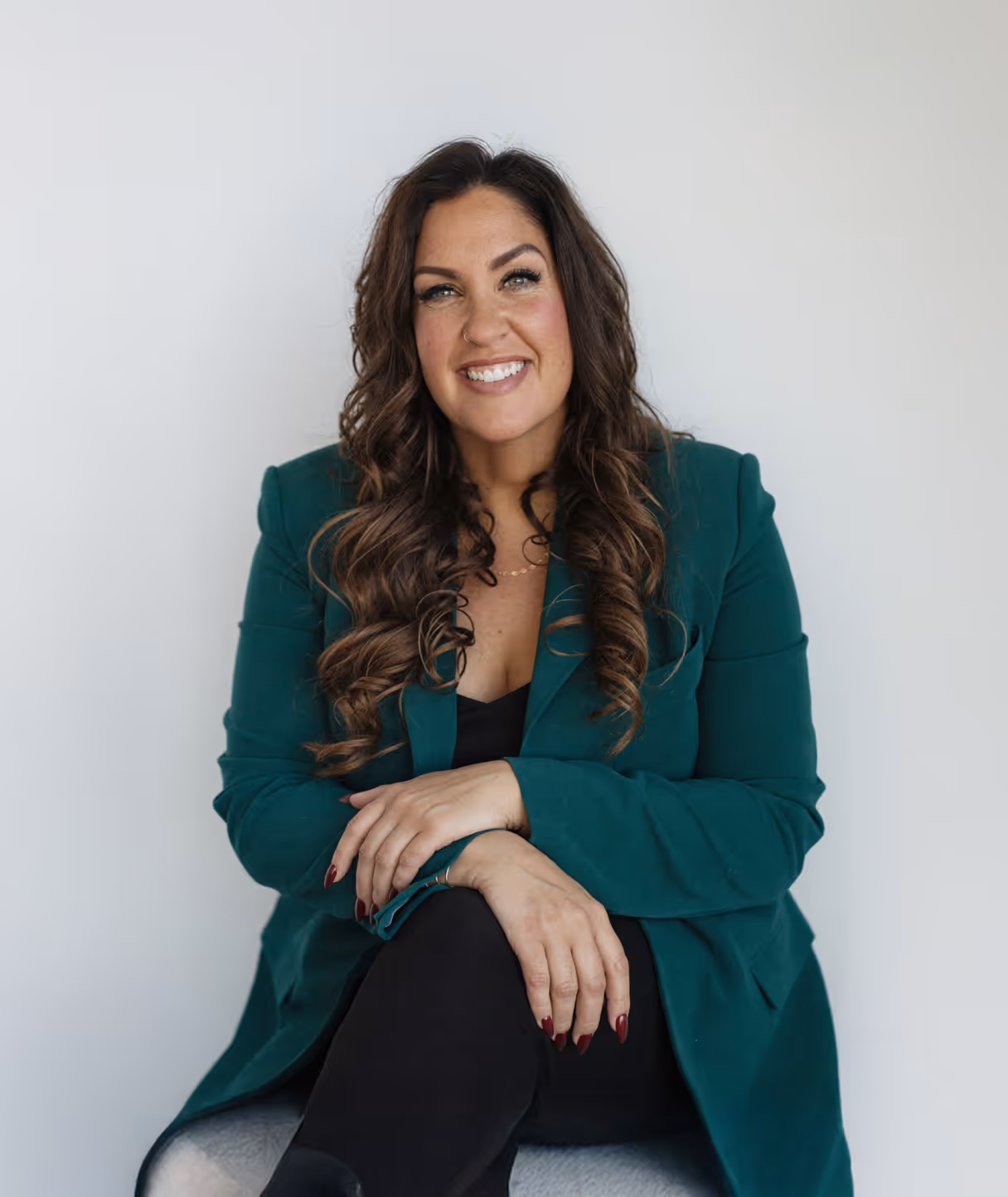 Smiling woman with long curly brown hair wearing a green blazer and black outfit, seated against a plain light background.
