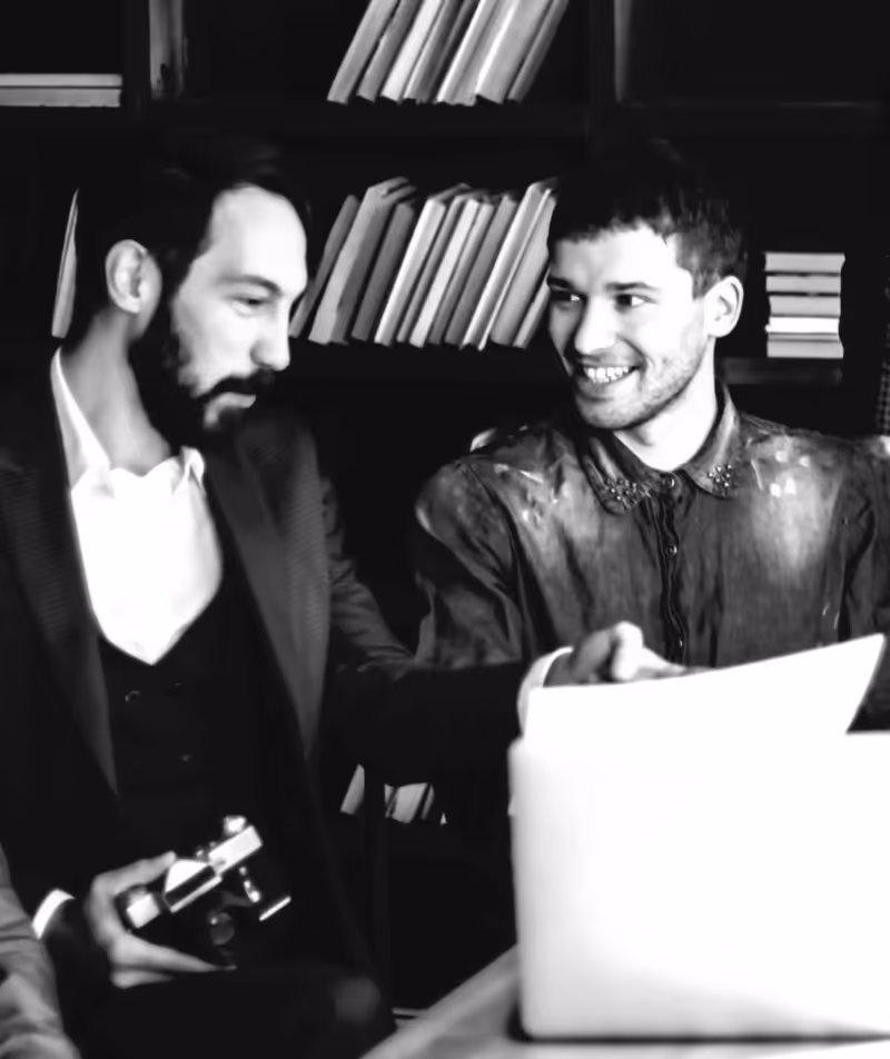 Two men sitting at a table with a laptop, one holding a camera, smiling and talking in front of bookshelves.