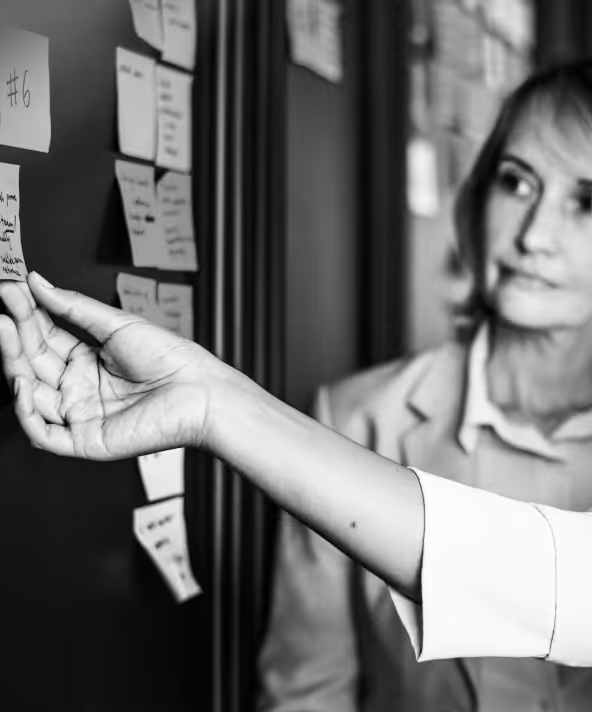 Person pointing at sticky notes arranged on a wall while a woman in business attire looks on attentively.