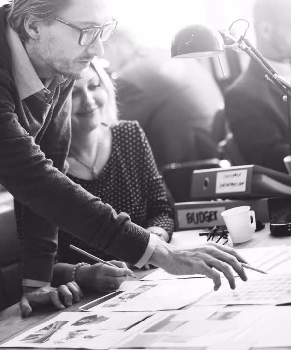 Man and woman collaborating over documents at a table with office supplies and a lamp.
