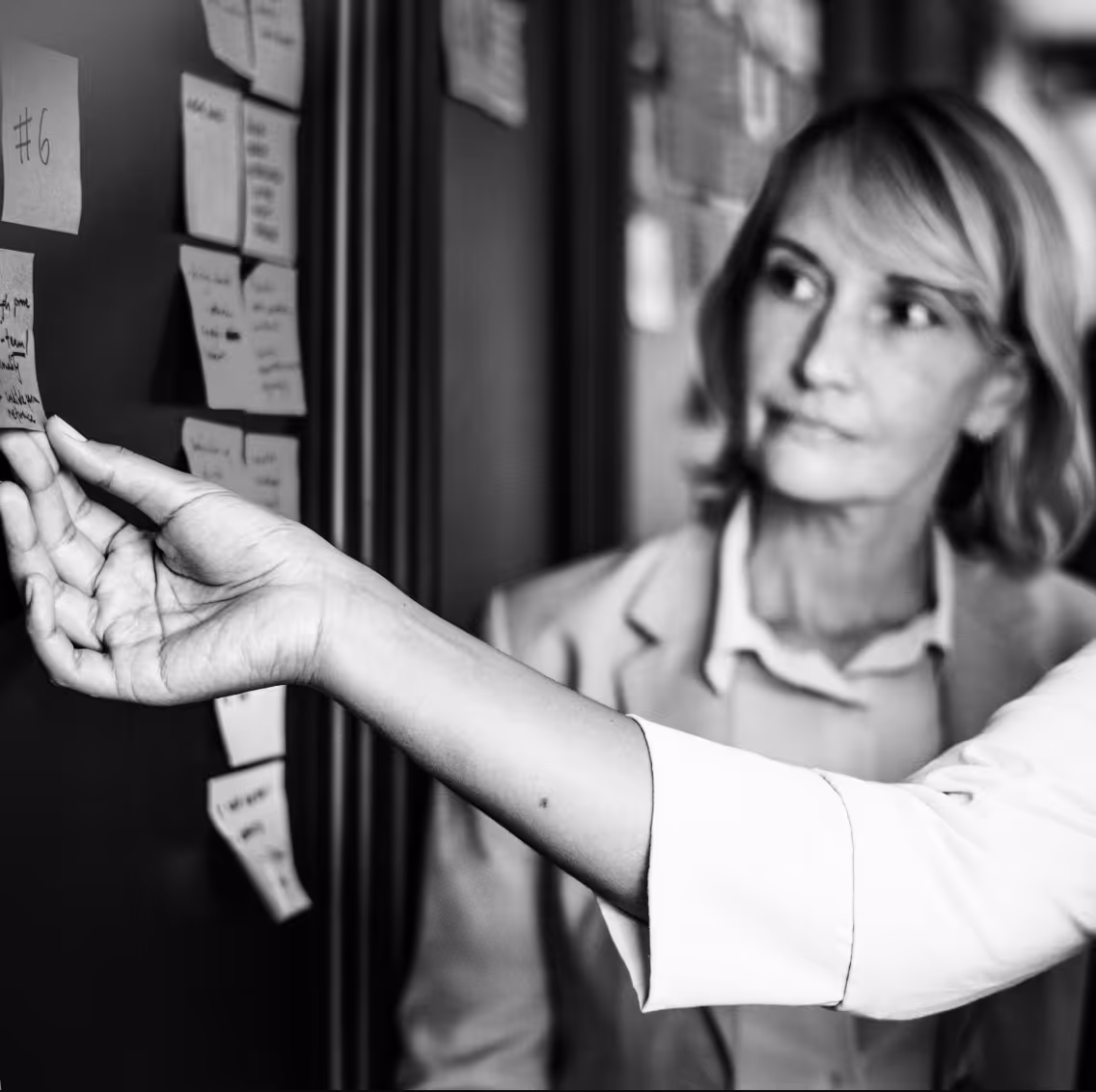 Woman in a blazer attentively looking at a hand placing sticky notes on a wall.