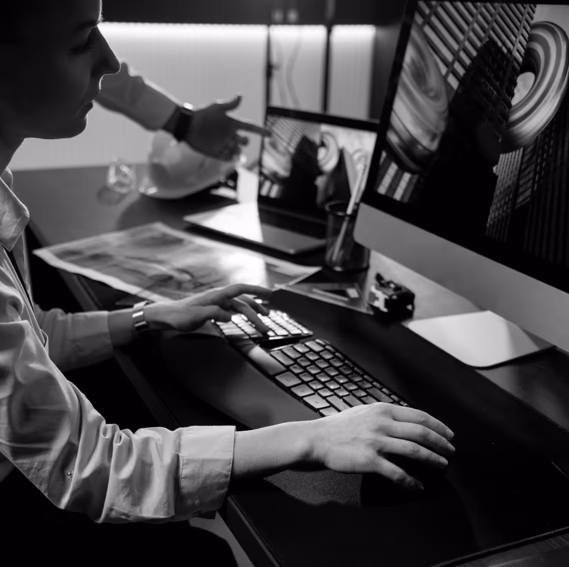 Person using a computer mouse and keyboard while another person points at a screen displaying curved architectural images in an office setting.