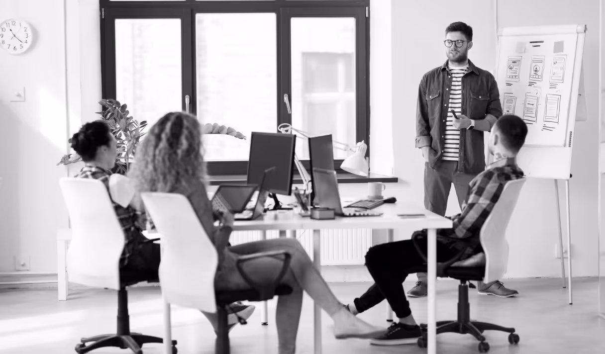 Man standing next to a flip chart presenting to three seated coworkers in a modern office.