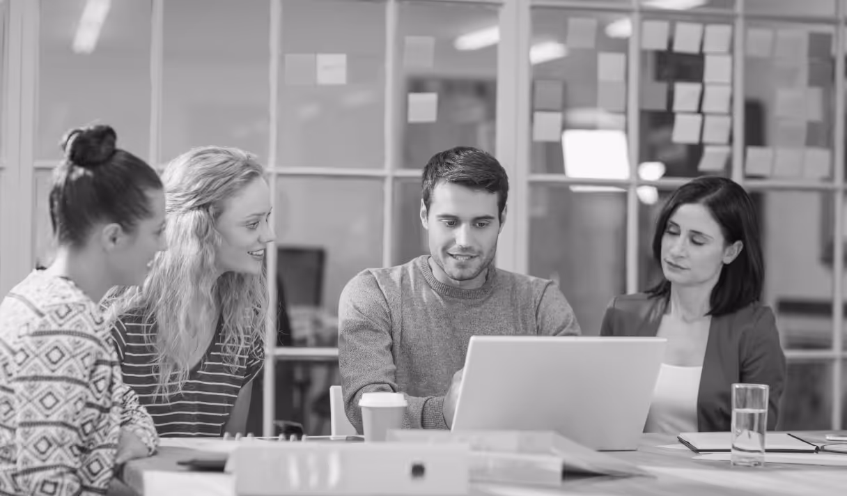 Four colleagues sitting at a table in an office, looking at a laptop during a meeting.