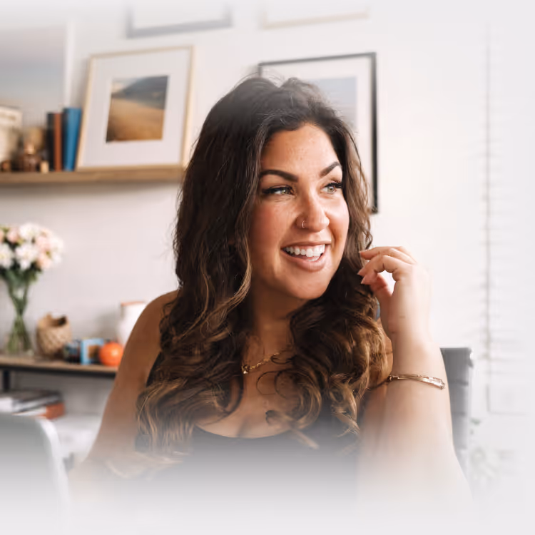 Smiling woman with long curly hair sitting in a room with framed pictures and flowers in the background.