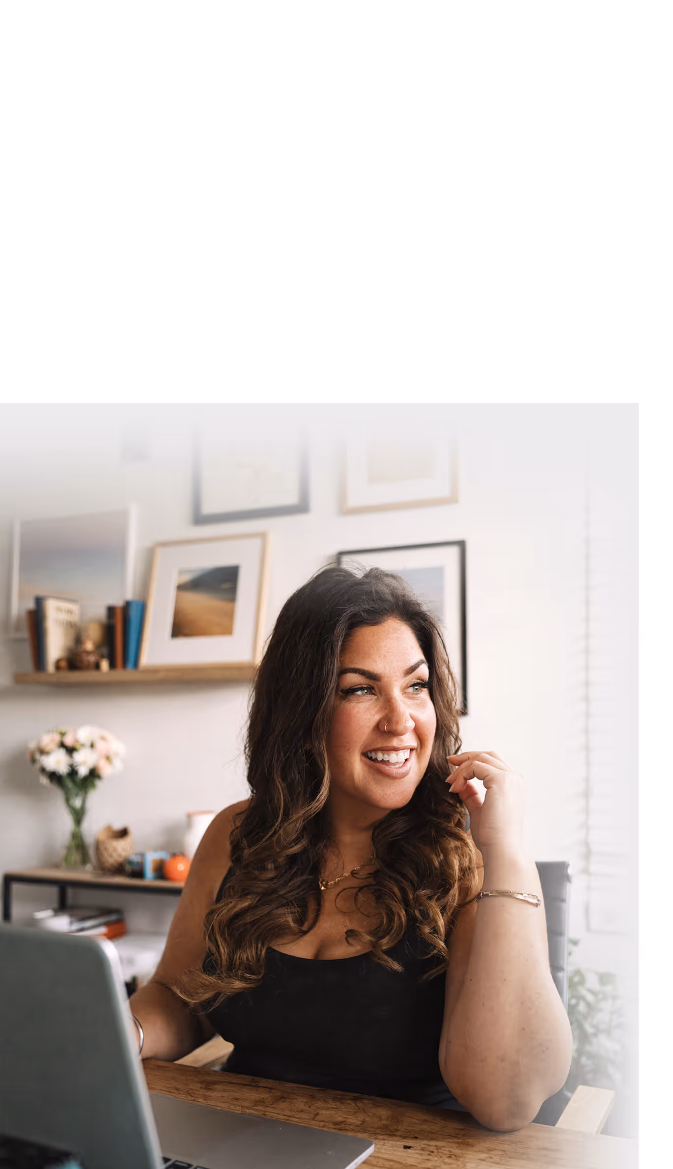 Smiling woman with long curly hair sitting at a wooden desk with a laptop, in a cozy room with framed pictures and flowers in the background.