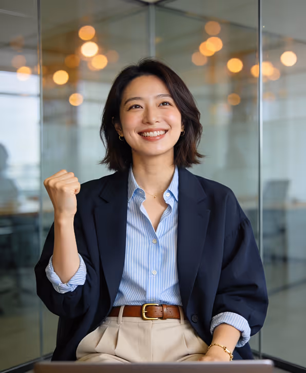 Smiling woman in business attire raising a clenched fist in a glass-walled office.