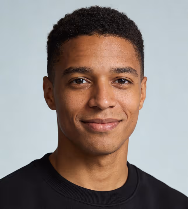 Portrait of a young man with short curly hair wearing a black shirt, smiling slightly against a light gray background.