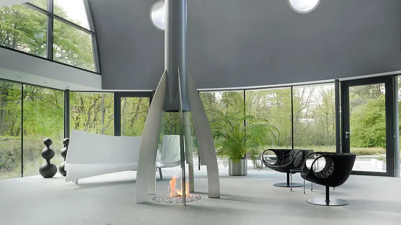 Modern room with tall glass fireplace, black chairs, white sofa, and large windows showing green trees.