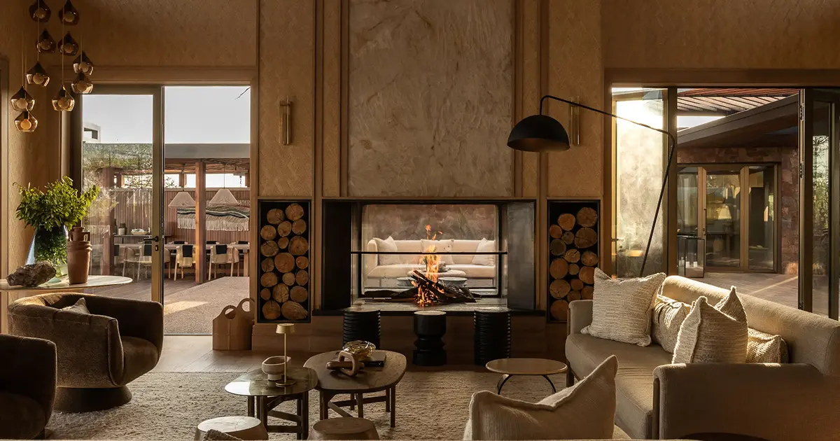Cozy living room with a central fireplace, sofas, armchair, and outdoor dining visible through glass doors.