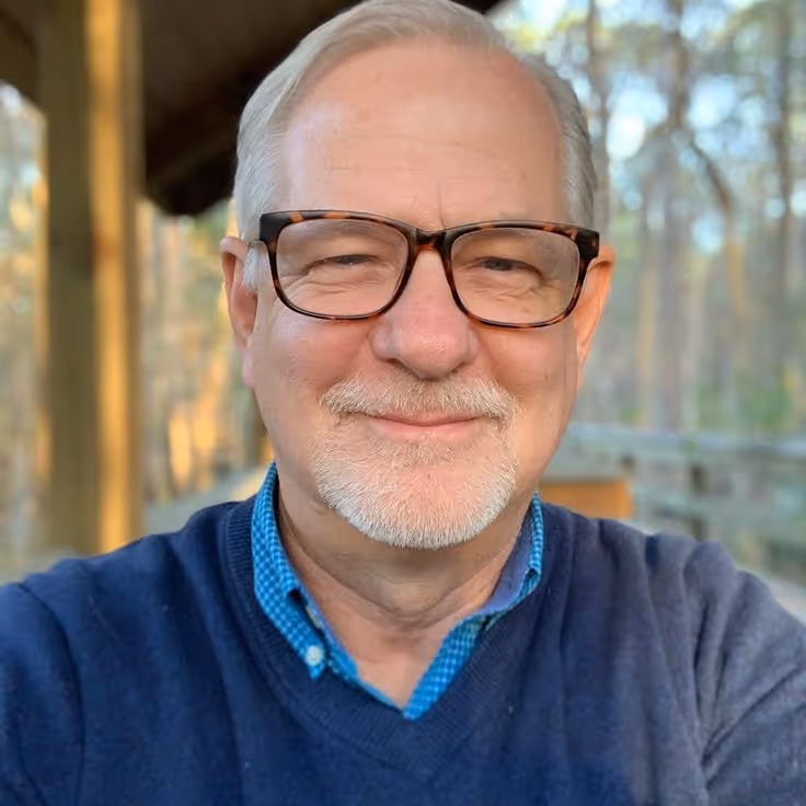 Smiling elderly man with white hair, glasses, a gray sweater, and blue checkered shirt outdoors with trees in the background.