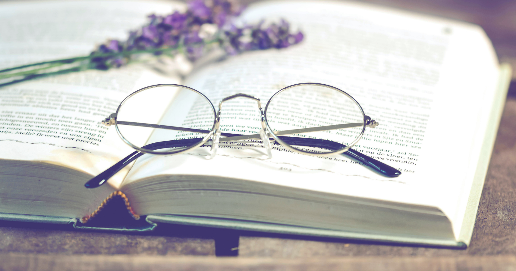 Round glasses resting on an open book with purple flowers in the background.