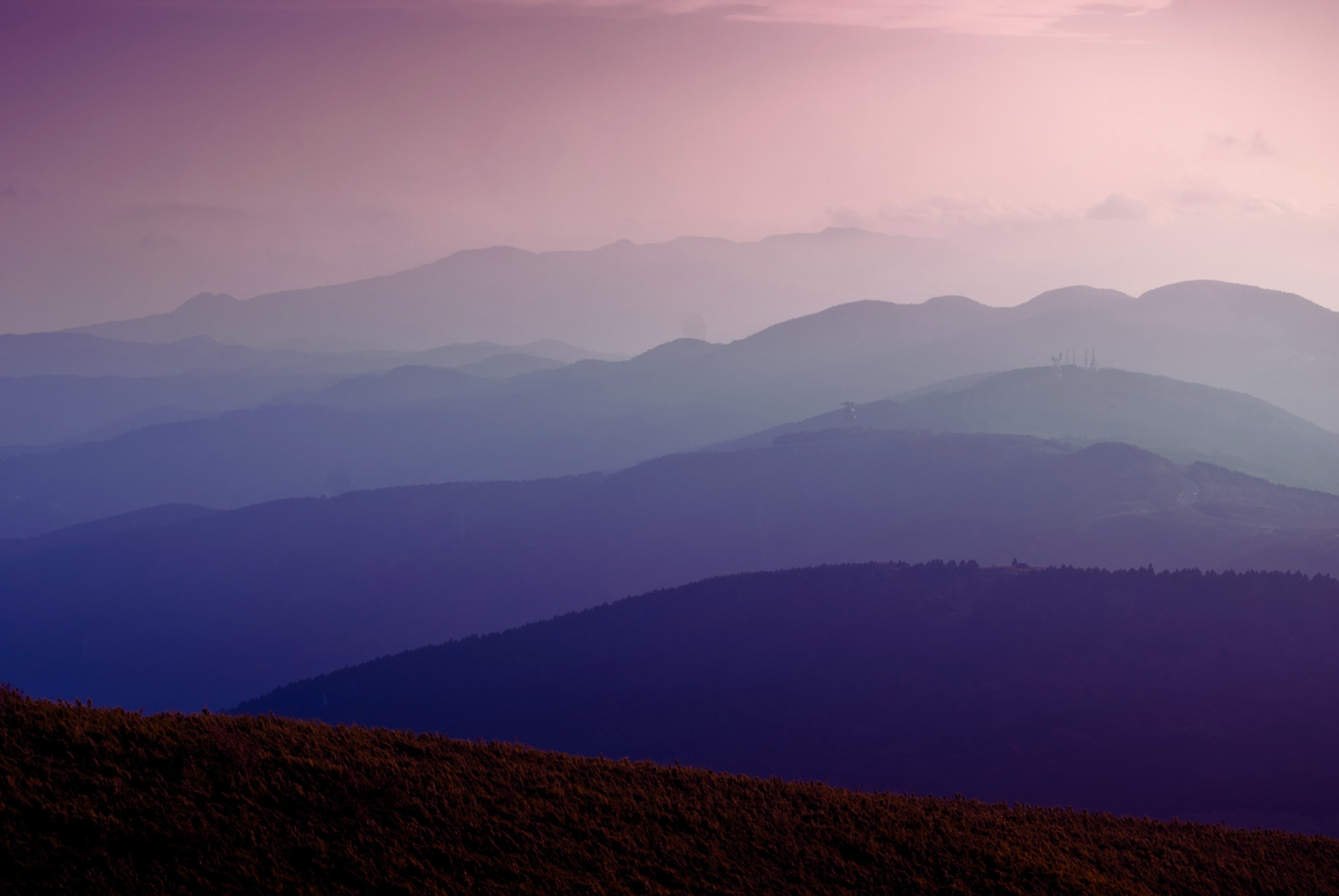 Layered mountain ranges in purple and pink hues under a soft, cloudy sky at dawn or dusk.