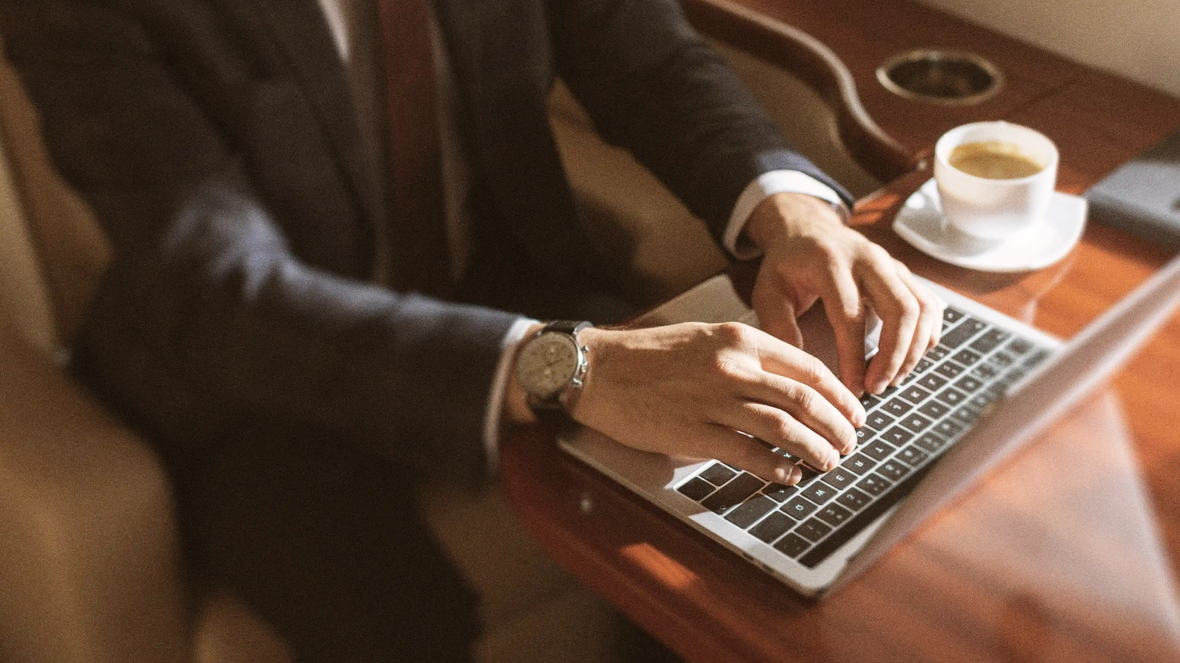 Person in a suit typing on a laptop at a wooden table with a cup of coffee nearby.