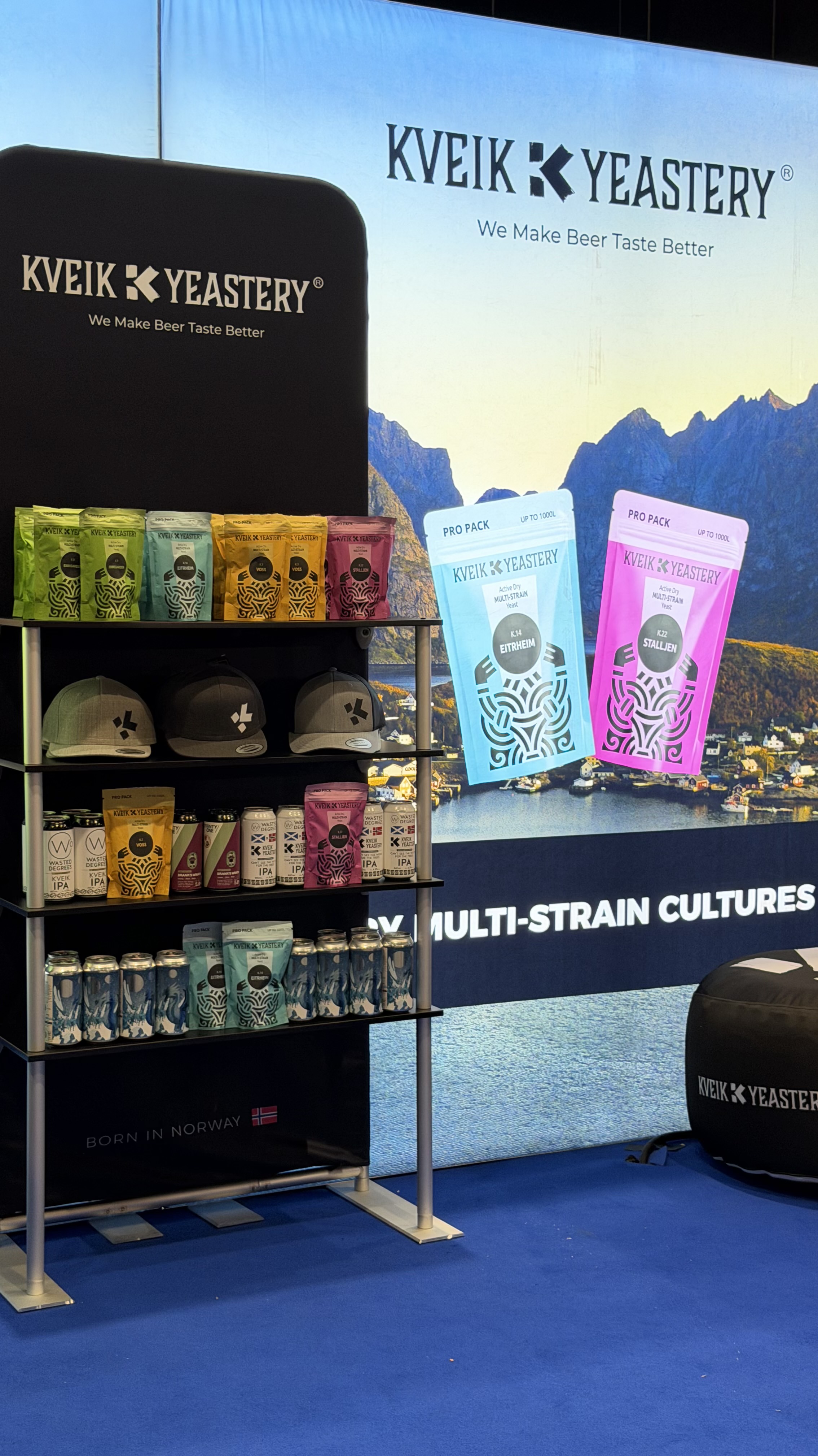 Shelf display of Kveik Yeastery beer yeast packs, branded caps, and beer cans at a trade booth.