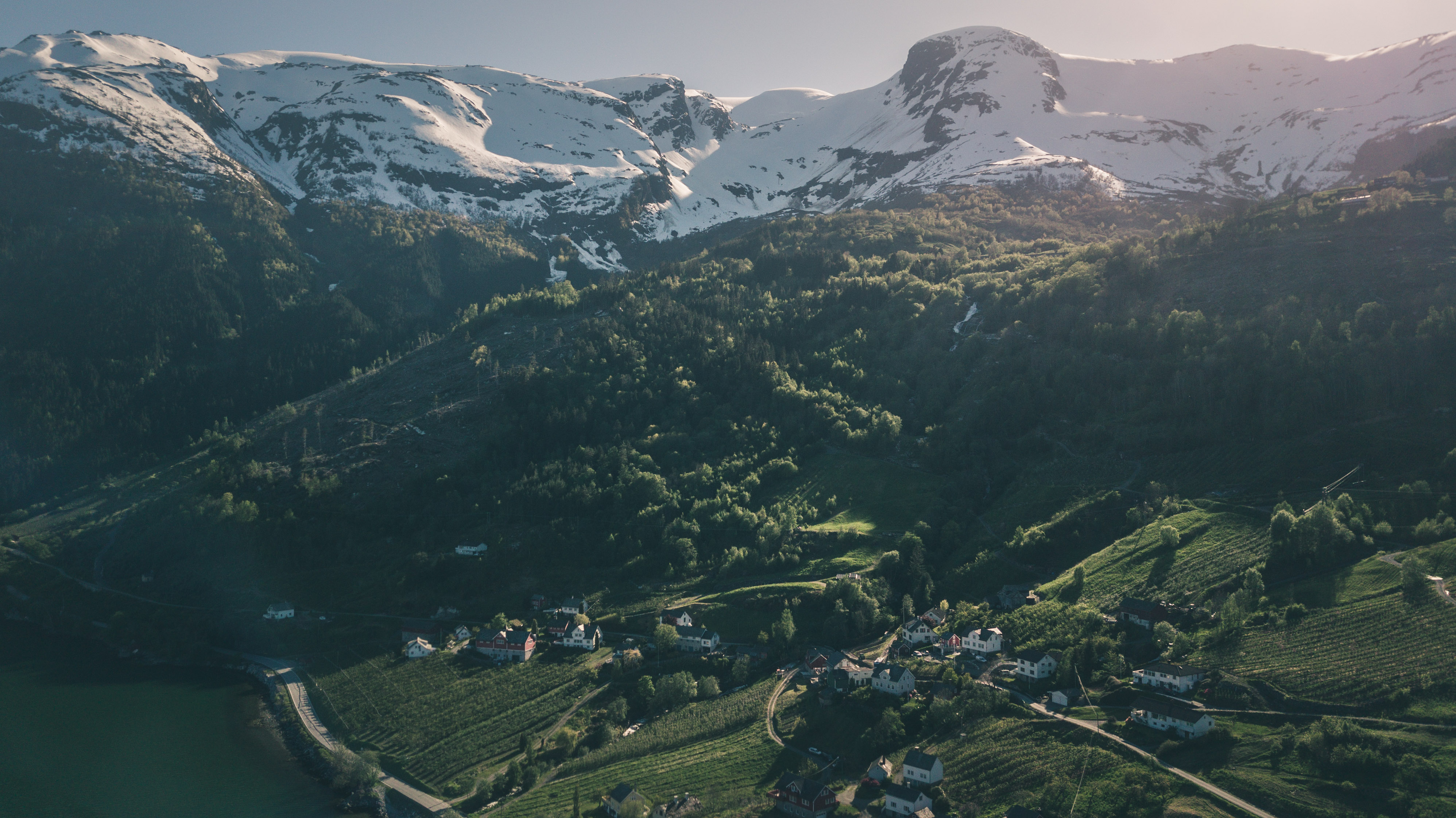 Small village with houses and green fields below snow-capped mountains under soft sunlight.