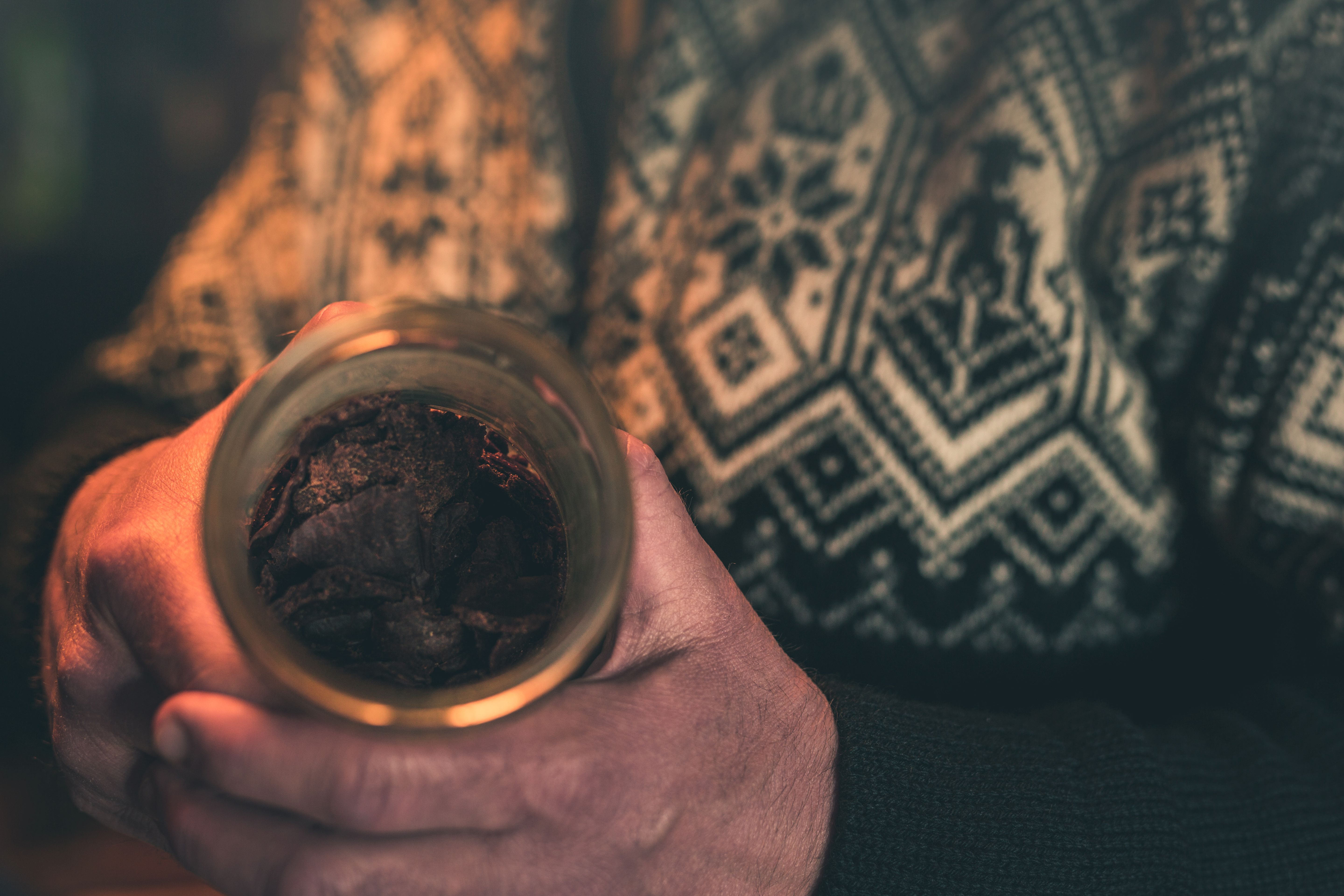 Hands holding a glass jar containing dark dried leaves, wearing a patterned sweater.