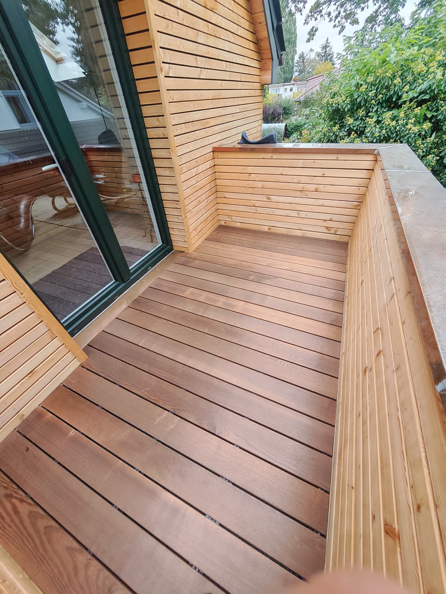 Holzterrasse mit Holzgeländer und grüner Gabeltür, umgeben von Bäumen und Häusern.