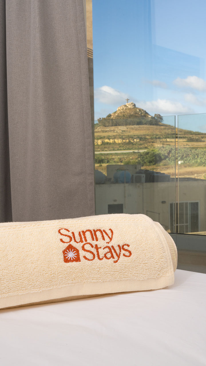 Rolled Sunny Stays towel on a bed, with the Gozitan view in the background.