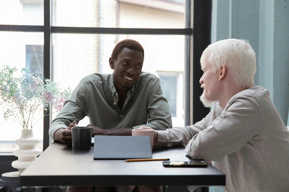 Two men sitting at a table by a window, talking and holding coffee mugs in a relaxed setting.