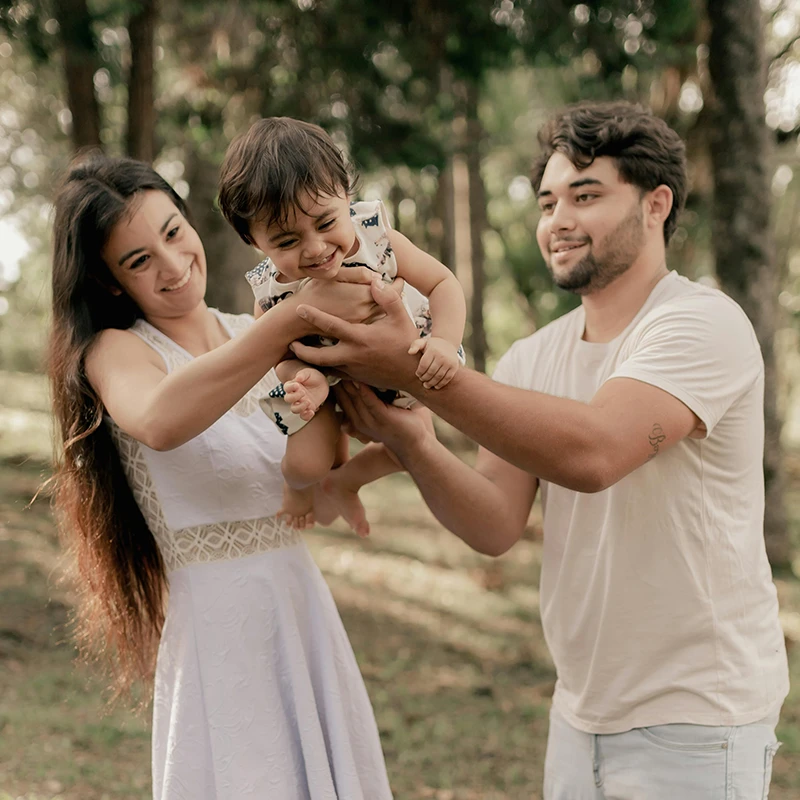 Man and woman holding a laughing baby together outdoors among trees.