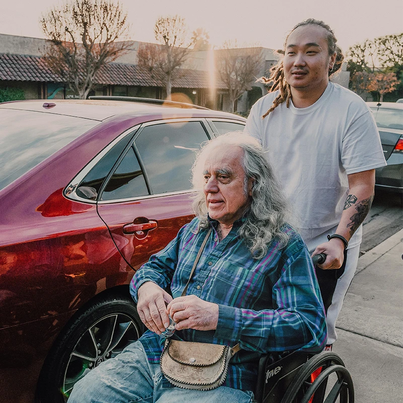 Younger person with tattoos pushing an older man with long gray hair in a wheelchair outdoors.