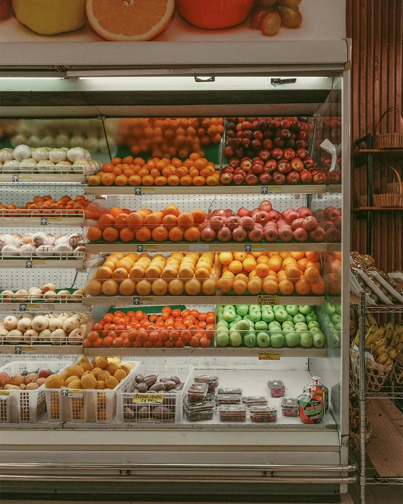 Grocery store refrigerated shelf with oranges, apples, garlic, tomatoes, pears, and packaged berries.
