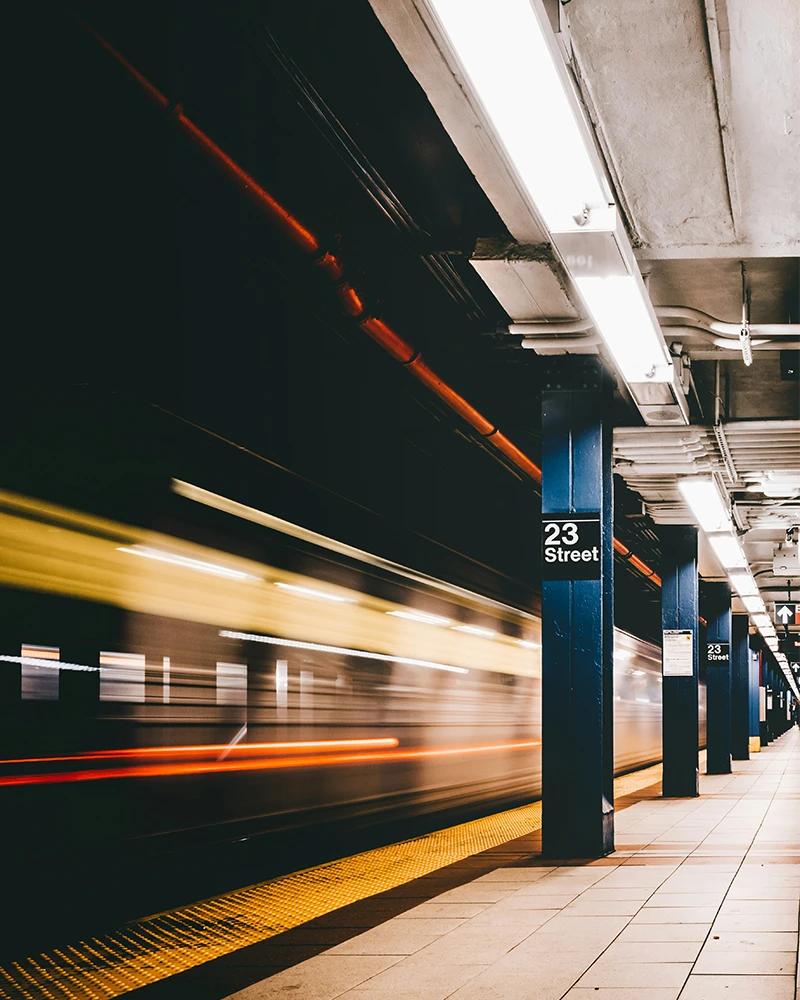 Blurred subway train speeding past 23 Street station platform with blue pillars and tiled floor.