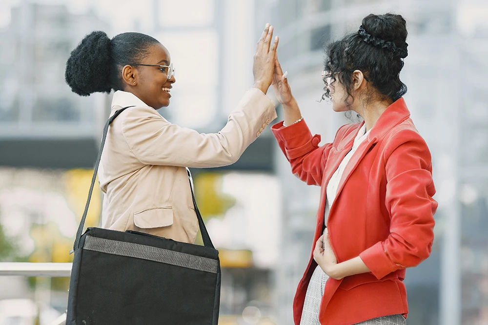 Two women smiling and giving each other a high five outdoors in a business district.