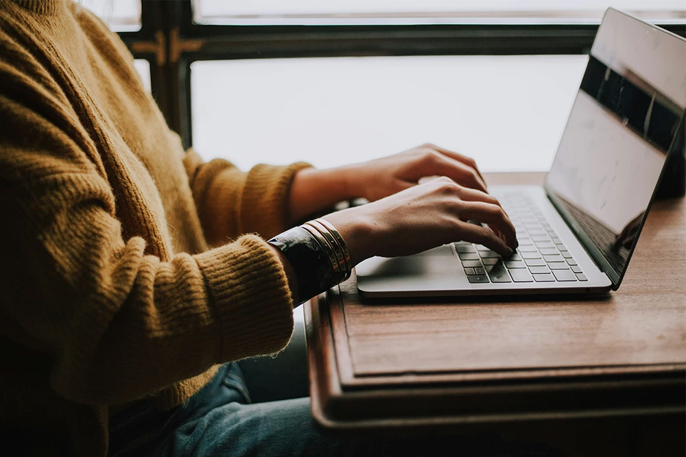 Person in a brown sweater typing on a laptop at a wooden desk near a window.
