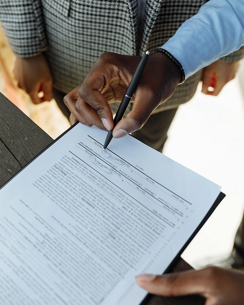Person signing a detailed contract document with a black pen held by a hand wearing a blue sleeve.