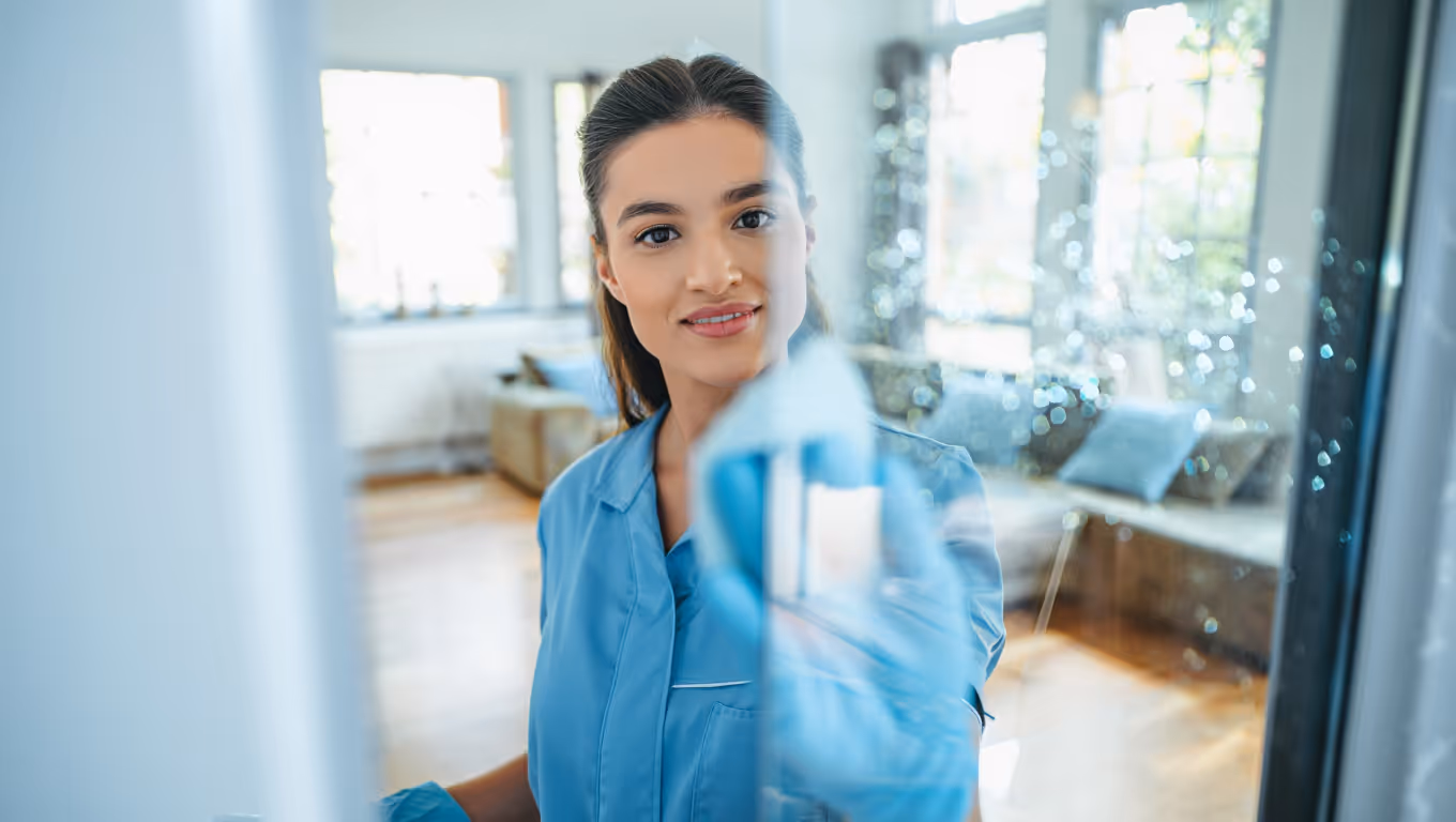 Femme en uniforme bleu nettoyant une vitre avec un chiffon dans un salon lumineux.