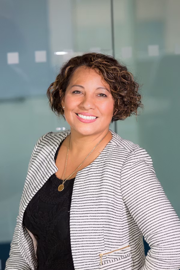 Femme souriante aux cheveux bouclés courts portant un blazer rayé blanc et noir et un pendentif doré.