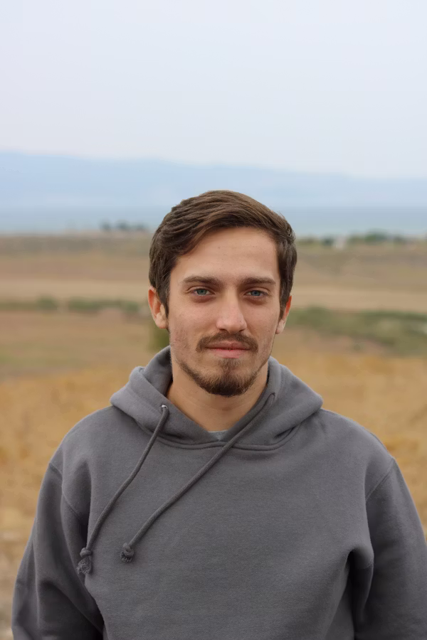 Jeune homme aux cheveux bruns portant un sweat à capuche gris dans un paysage ouvert flou.