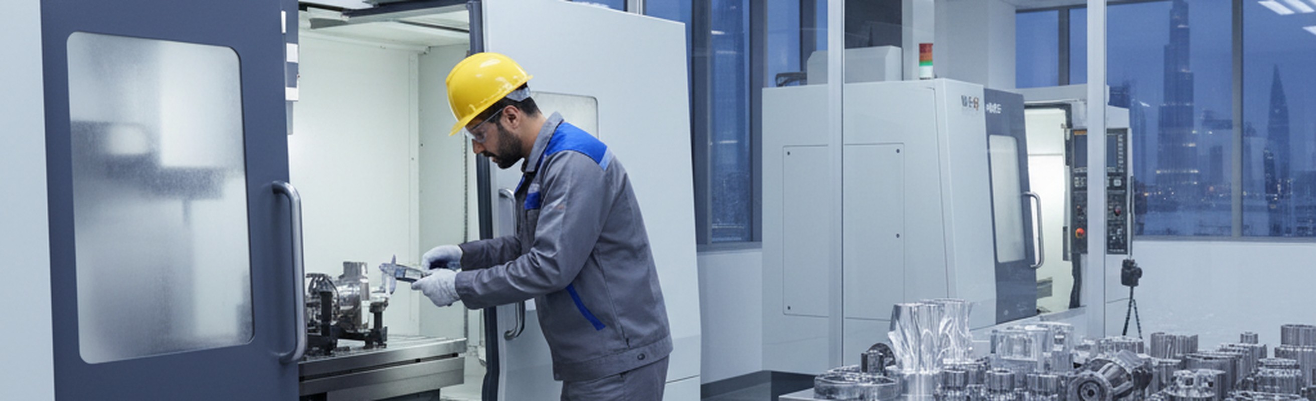 Machinist in a yellow hard hat inspecting a component beside CNC machines in a UAE manufacturing facility