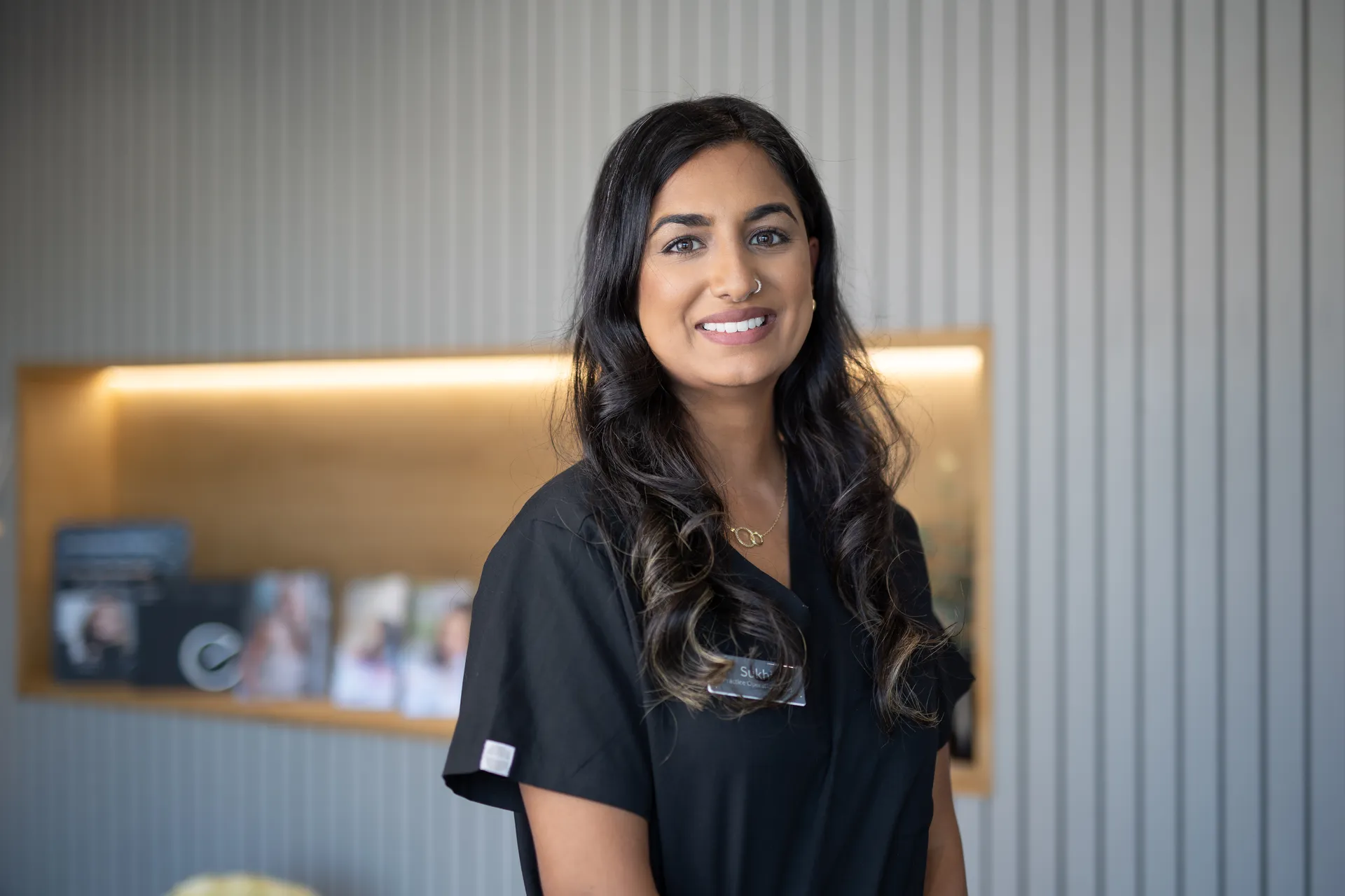 Smiling woman with long dark hair in black scrub top standing against a gray striped wall.
