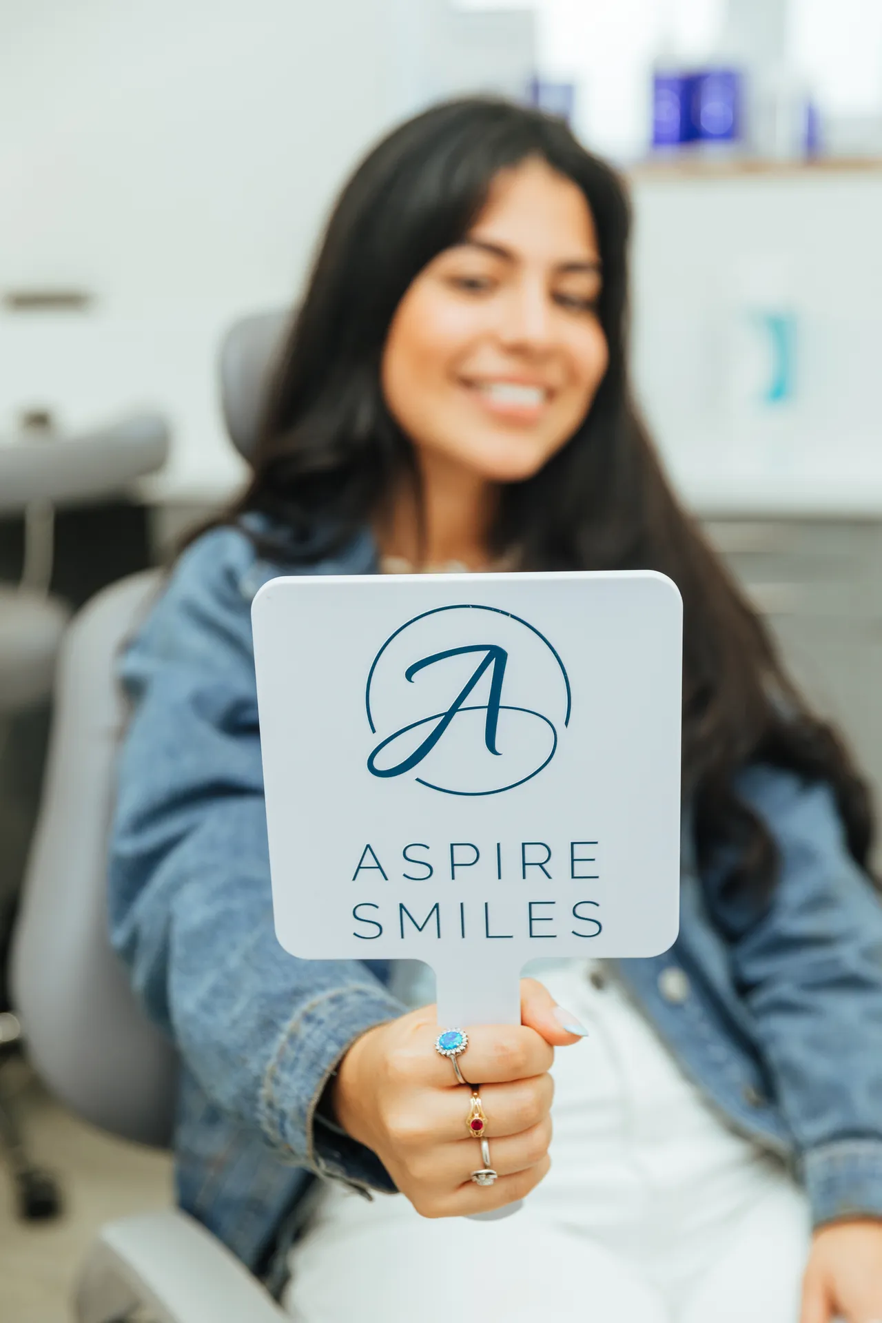 Woman holding a mirror with Aspire Smiles logo, smiling in a dental office chair.