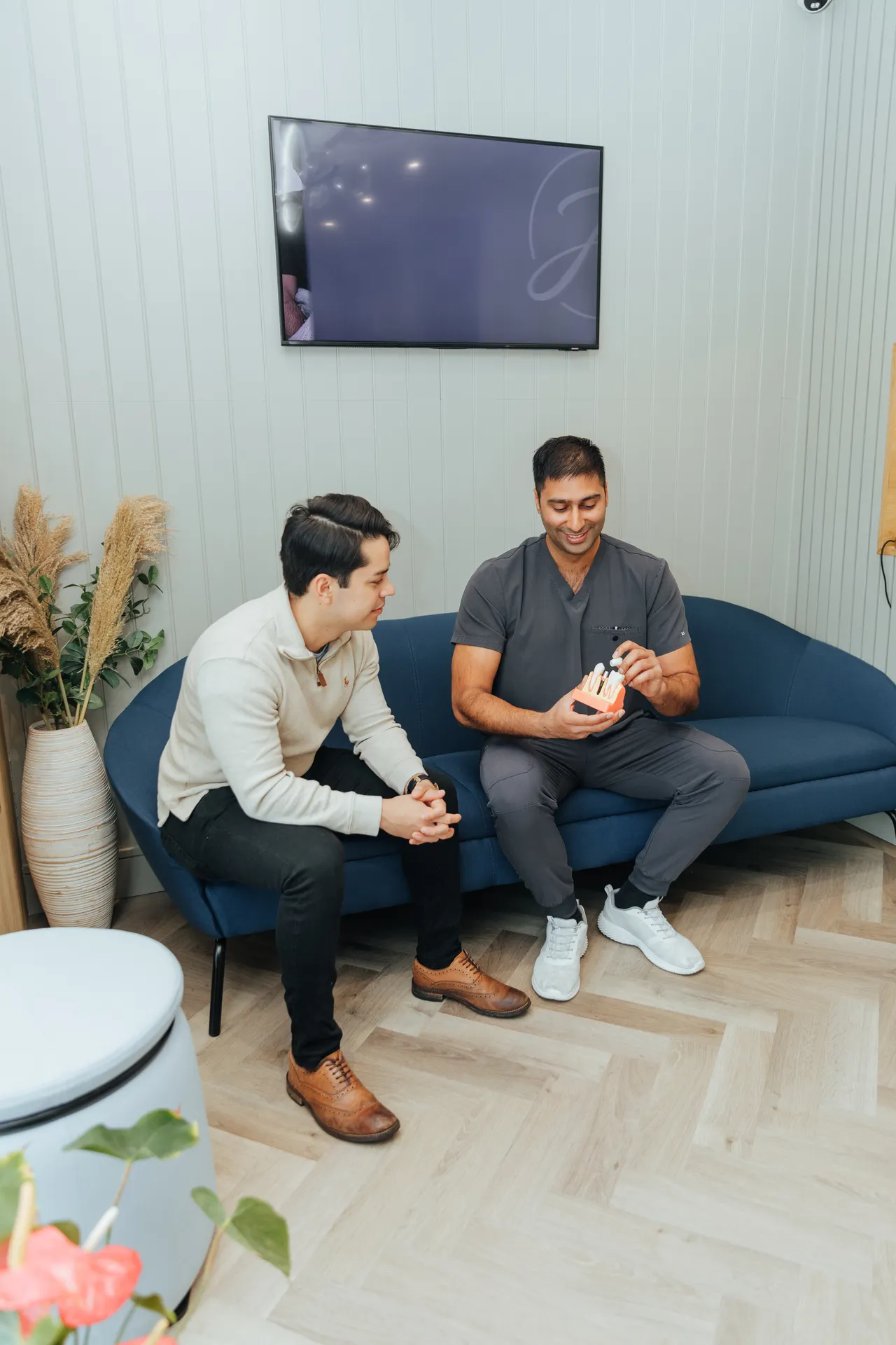 Man in scrubs shows dental model to a seated man in a white sweater on a blue couch.