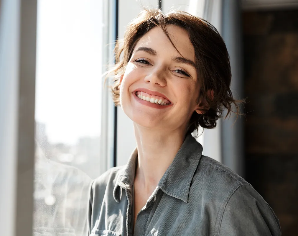 Young woman with short hair smiling brightly in natural light by a window.