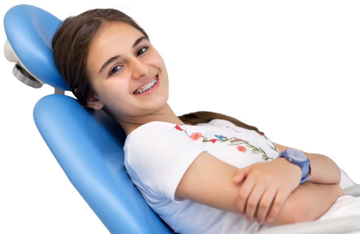 Smiling teenage girl with braces reclining in a blue dental chair wearing a white shirt with floral embroidery.