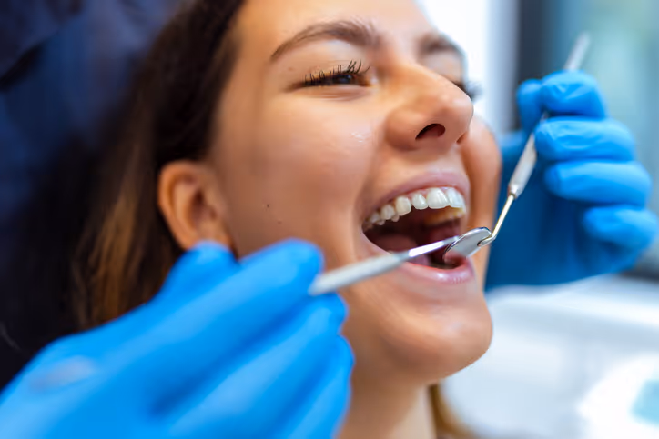 Young woman with braces smiling during a dental checkup with a dentist wearing blue gloves holding dental tools in her mouth.