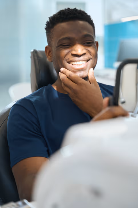 Smiling man sitting in a chair looking into a small mirror while touching his chin.