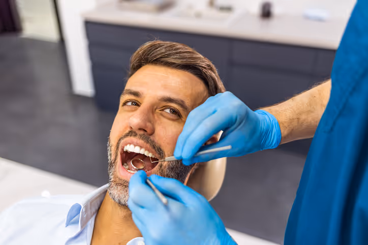 Man with beard receiving dental checkup from a dentist wearing blue gloves and scrubs.